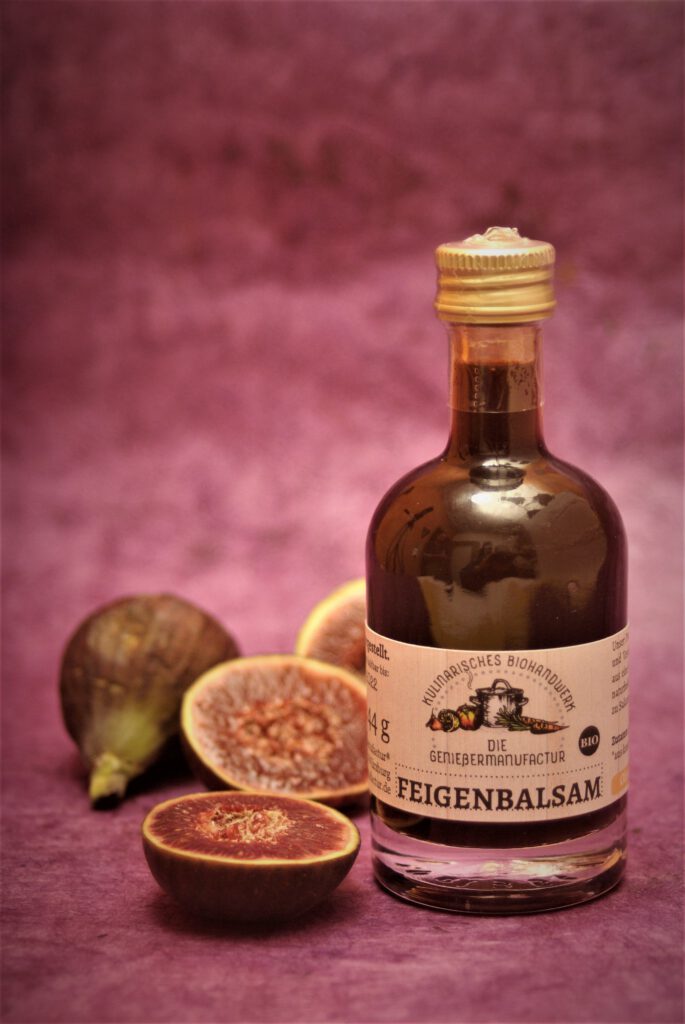 Produktfotografie Feigenbalsam von der Geniessermanufactur in der Flasche mit frischen Feigen auf lilafarbenem Hintergrund.